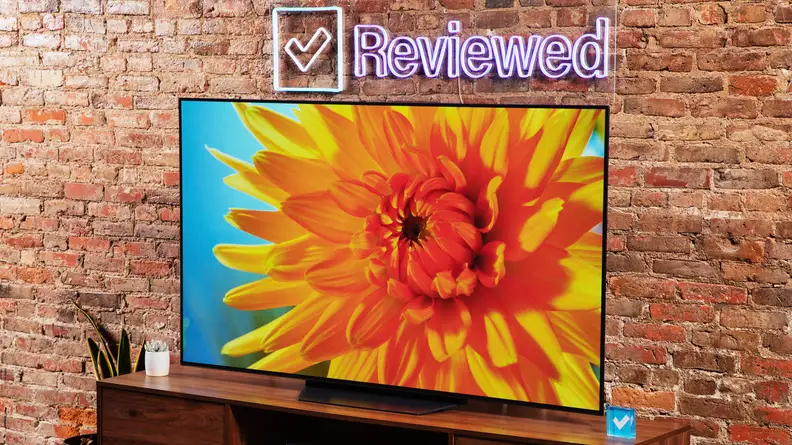 The LG B2 on a wooden table in front of a brick wall with a neon Reviewed sign displaying a flower.