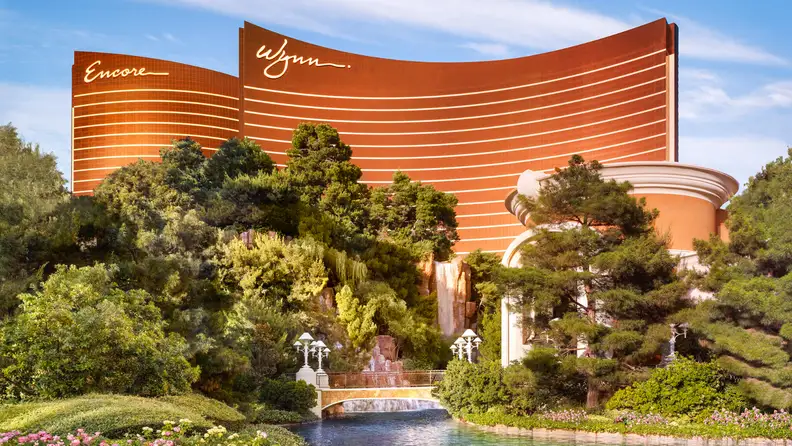Wynn and Encore Las Vegas towers rising above lush landscaped gardens, waterfalls, and reflective water features in the foreground.