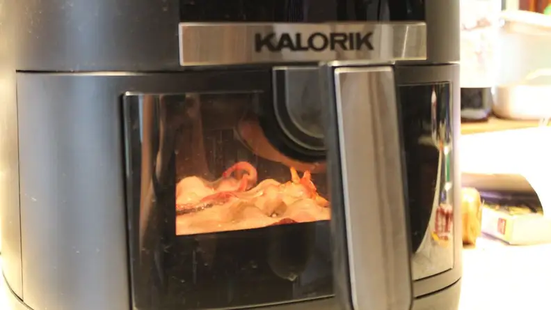 Bacon being cooked inside of basket while seen through the small window behind handle of the Kalorik 5-Quart Digital Air Fryer.