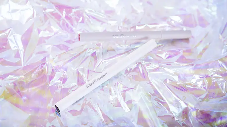A white tube of liquid eyeliner front and center with a gray tube of lip stain in the background, both on holographic cellophane paper.