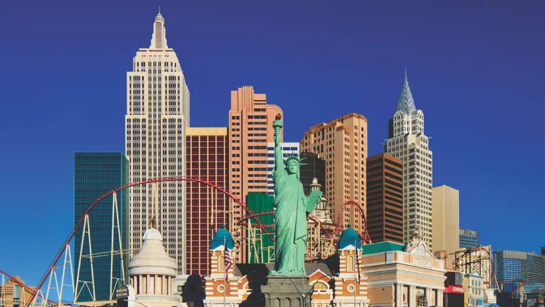 New York-New York Hotel & Casino showcasing a replica New York City skyline, Statue of Liberty, and roller coaster set against a clear blue sky.