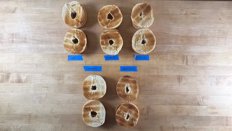 10 bagel halves with varying browning laid out on a countertop with distinguishing labels from respective toaster brands.