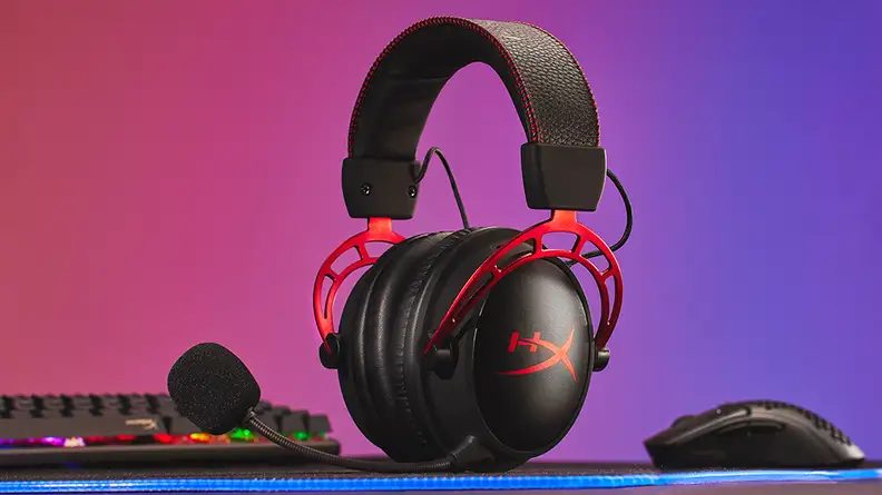A black gaming headset with red trim sits on a desk next to computing peripherals with a rainbow backdrop.