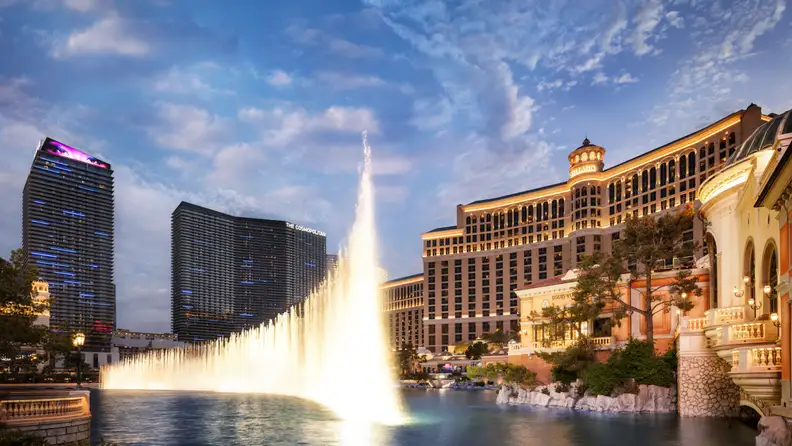A brightly lit casino hotel at dusk with fountains in front of it
