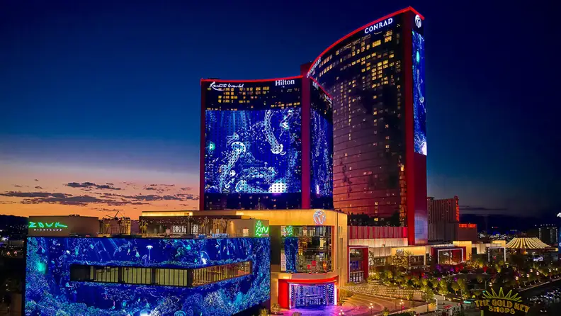 Resorts World Las Vegas illuminated at dusk, with vibrant digital displays covering the hotel towers and entertainment complex against a twilight sky.