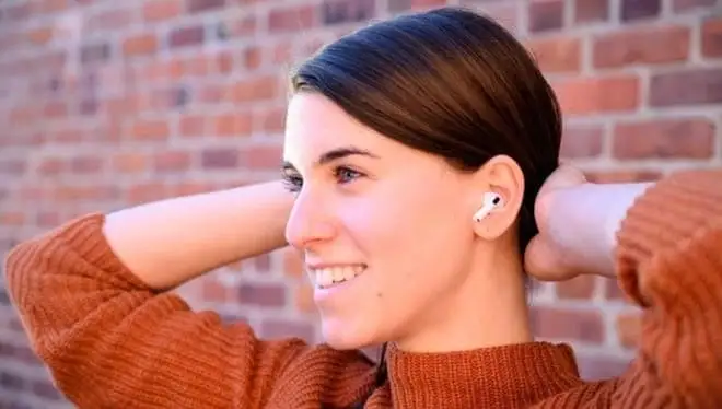 A woman sitting back in an orange shirt in front of a brick background wearing a pair of AirPods Pro.