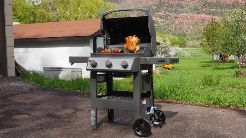 A Weber grill opened, revealing a whole chicken and veggie skewers, set in a backyard.