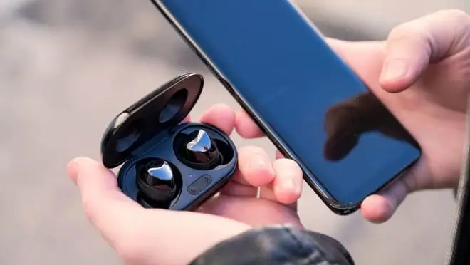 A black touchscreen phone in one hand, and black earbuds in the other