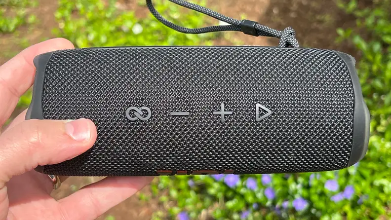 A black, cylindrical Bluetooth speaker is held above some flowers with its buttons facing outward.