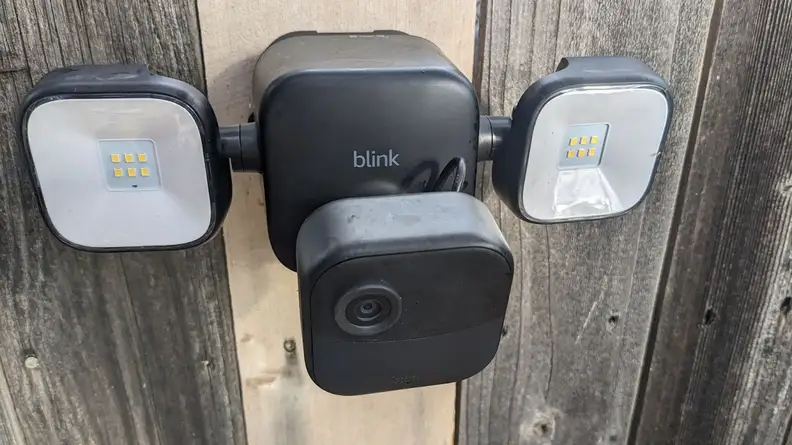 Downward aerial view of the Blink Outdoor 4 Floodlight Camera mounted on wooden fence outdoors with two identical floodlights located on both sides.