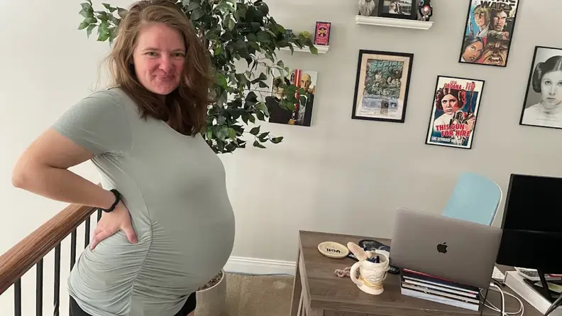 Pregnant person posing with hand on hip next to desk while wearing a Seraphine maternity top.
