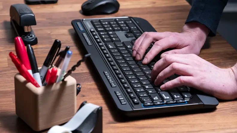 A black wireless keyboard sits on a wooden desktop; a wireless mouse, a stapler, a pen holder, and a tape dispenser surround it. Two hands are typing on the keyboard.