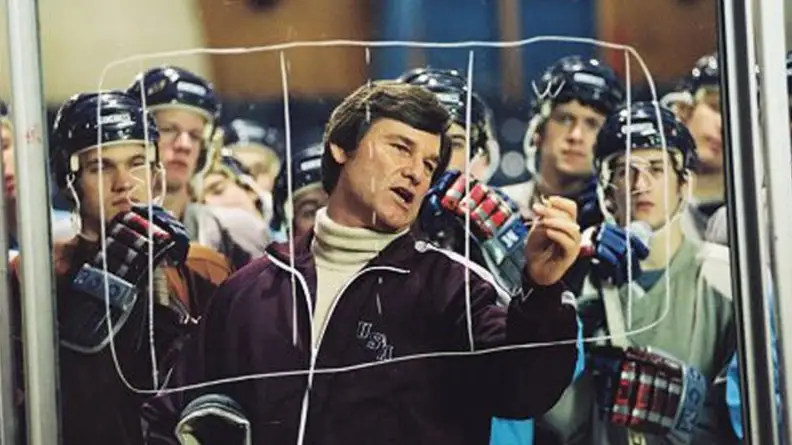 Actor Kurt Russel drawing a hockey play on a transparant board in front of his team of players.