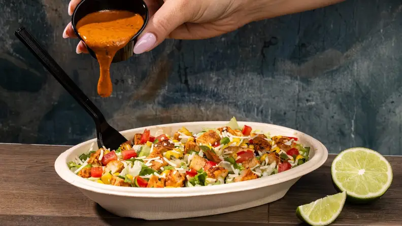 A hand pouring sauce over a chicken salad bowl with lime wedges