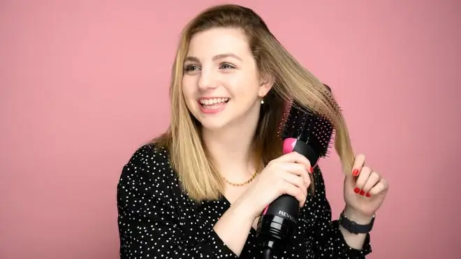 A woman standing in front of a pink background smiling and using a Revlon hair tool.