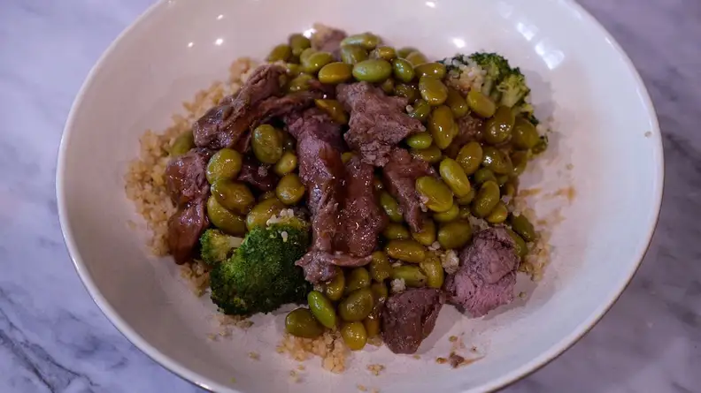 Cooked meat and vegetables inside of white bowl on countertop surface.