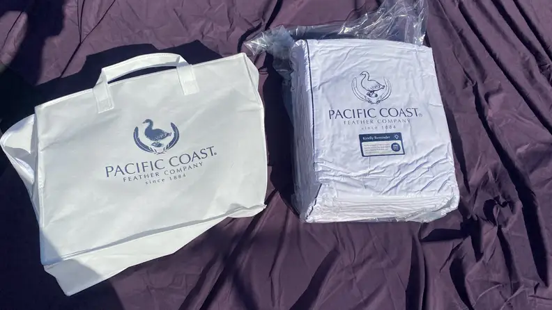 Packaging for a white Pacific Coast Feather & Down Comforter on top of a dark purple sheet.