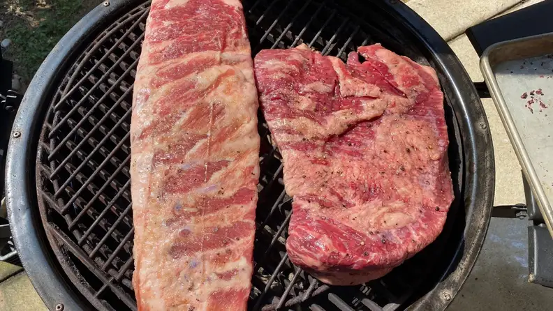 An uncooked rack of ribs and a roast sit on the grate of a charcoal grill, ready to be smoked.