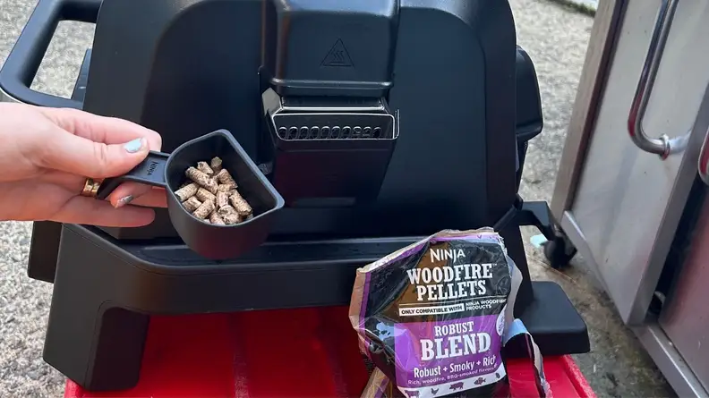 Person holds small black scooper filled with small amount of Woodfire pellets inside next to the side of the Ninja Woodfire ProConnect XL Outdoor Grill & Smoker and bag of Woodfire pellets.