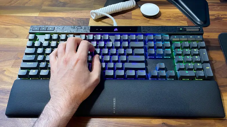 A left hand typing on a Corsair K70 Max RGB keyboard, with a wrist rest below it and full RGB on each key.
