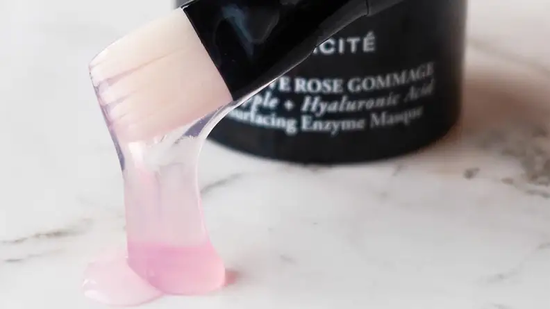 A closeup on a makeup brush with pink goop hanging from it. A face mask jar sits in the background.