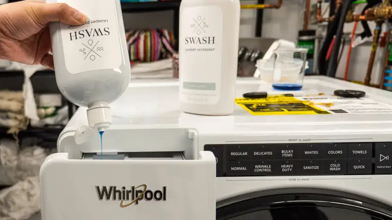 A hand reaches into frame holding a Swash bottle upside down, squeezing detergent into a washer.