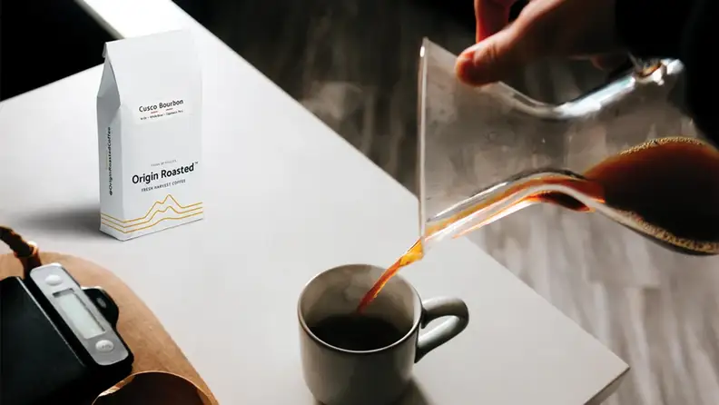 Coffee being poured from a glass carafe into a mug beside an Origin Roasted bag