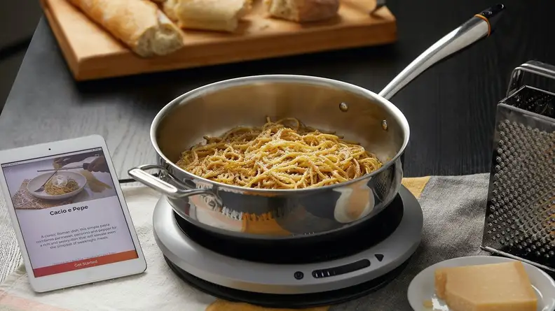 This smart induction cooktop walks you through each recipe, automatically adjusting its own temperature. Pasta with parmesan cheese is shown inside the pot; bread and a cheese grater are visible at the edges of the photo.