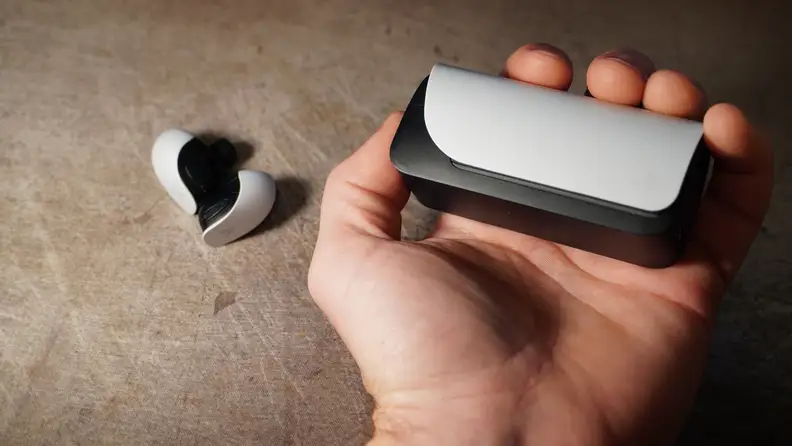 A hand holding the earbud case with the earbuds themselves laying on a table.