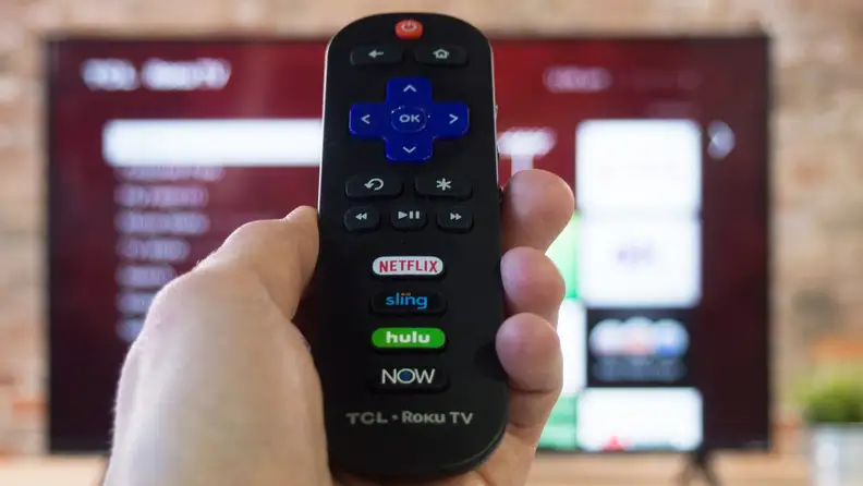 Person holding up Roku remote in front of flat screen smart TV.