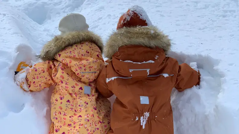 Two children laying down in the snow while wearing snowsuits.