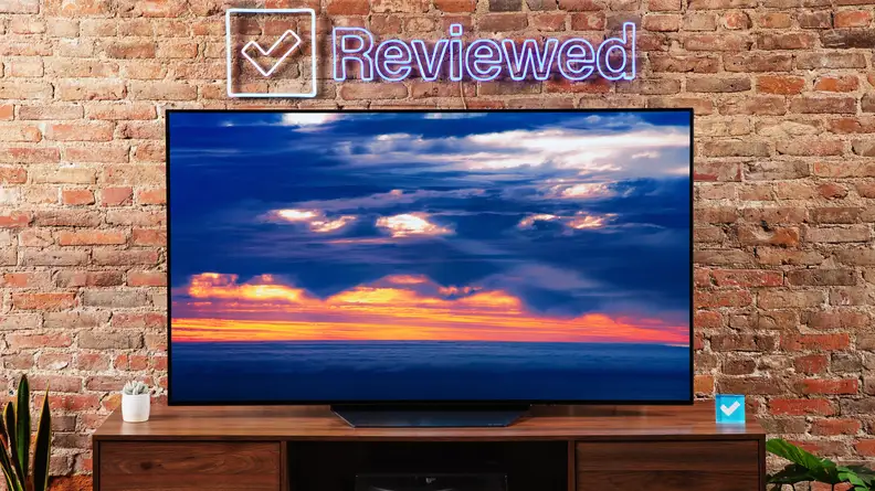 The LG B2 on a wooden table in front of a brick wall with a neon Reviewed sign displaying a sunset.