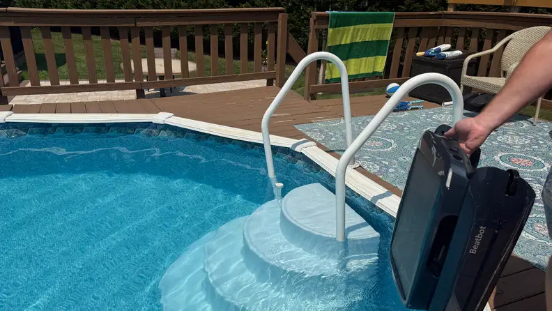 Black Beatbot iSkim being lowered into an above-ground pool with round steps on a brown deck.
