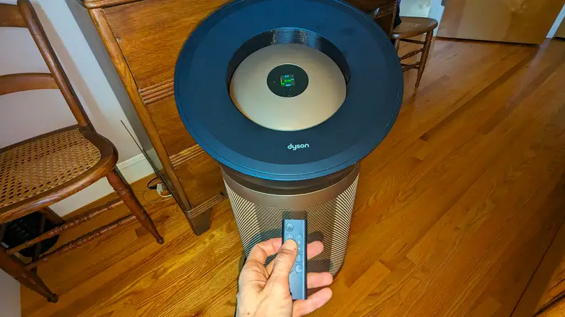 A hand holds a remote control and points it at the circular top of the Dyson air purifier.
