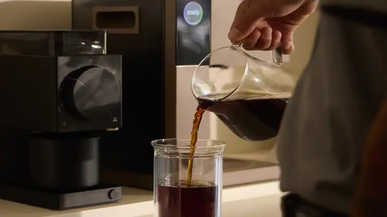 Hand pouring coffee from a glass carafe into a glass beside a Fellow grinder