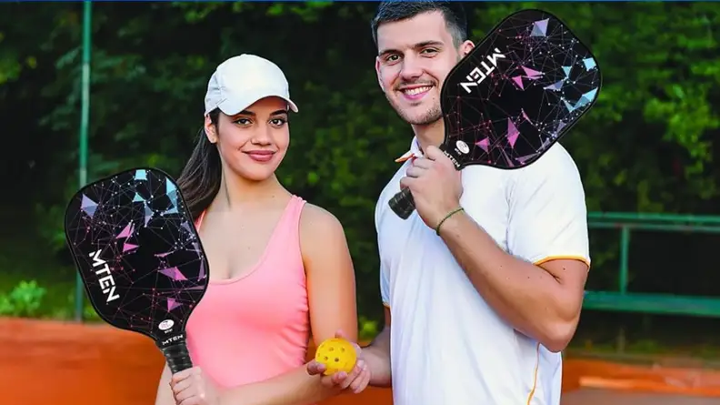 A woman and a man are holding pickleball paddles and balls on a court.
