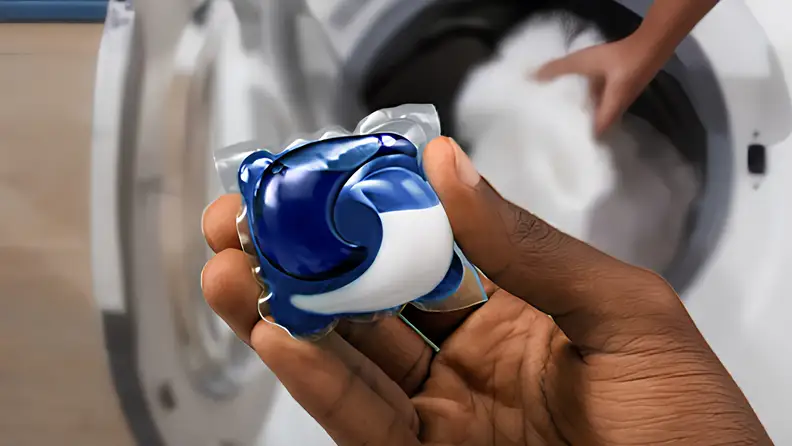 A hand holds a blue and white Tide laundry pod above an open front-load washing machine.