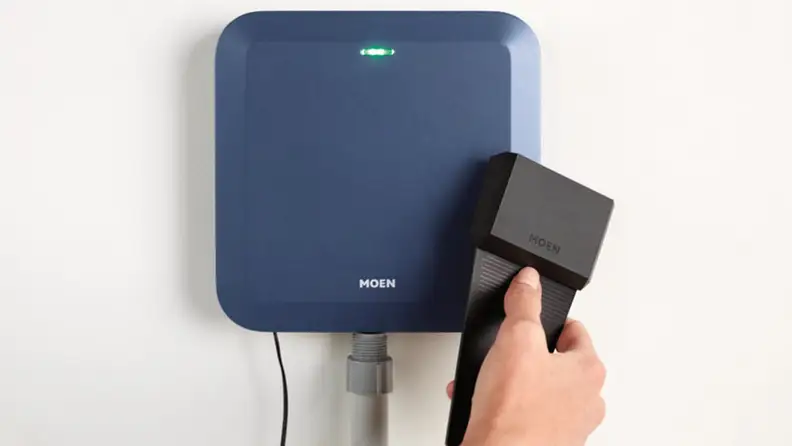 Person holding black soil sensor in front of Moen Smart Sprinkler Controller that’s mounted on a wall.