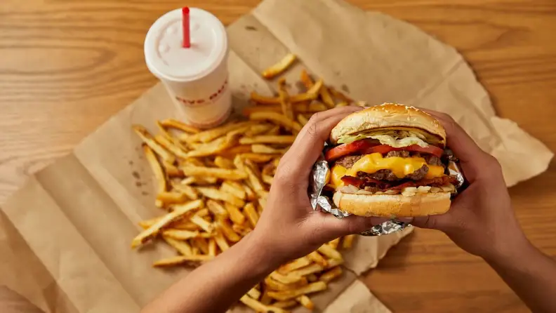 Hands holding a Five Guys burger with fries and a drink in the background