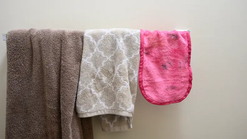The Makeup Eraser hung over a towel ring next to a hand towel and bath towel.