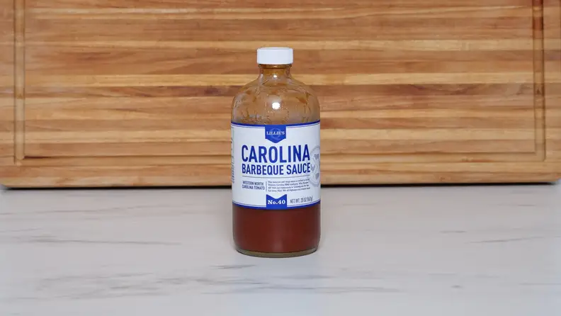 A bottle of Lillie’s Q Carolina Barbeque sauce next to a wood cutting board.