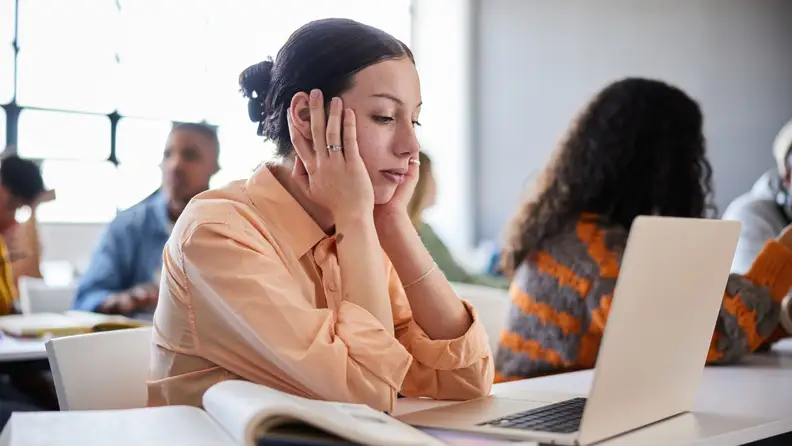 A person resting their face in their hands in front of a laptop computer in a classroom.