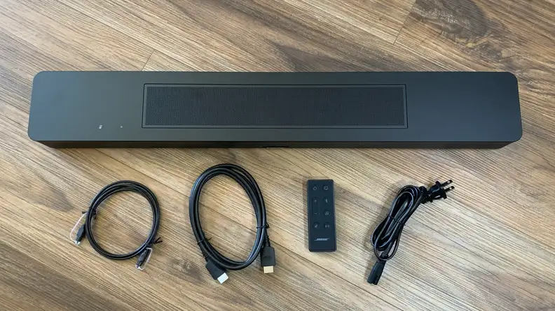 The Bose Smart Soundbar 600 sitting on a wooden floor with its remote, an optical, HDMI, and power cable.