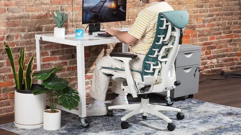 The side view of the Herman Miller x Logitech G Embody near a white desk, with a computer monitor and office supplies.