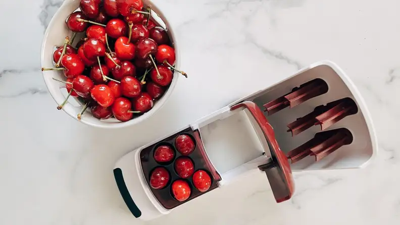 A bowl of cherries sits beside the OXO multi-cherry pitter on a marble surface.