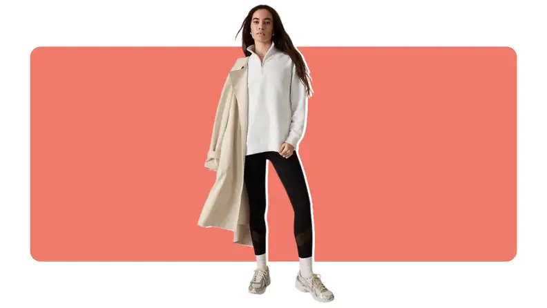 A model wearing black Salutation Stash high-rise leggings, a beige pullover and a stylish jacket resting on their shoulder with a matching pair of sneakers.