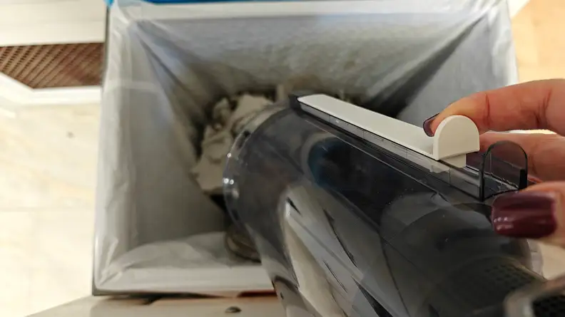 A hand empties a vacuum dust bin into a trash can