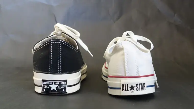 On the left, a Chuck 70 low top sneaker in black. On the right, a white Chuck Taylor All Star low top sneaker. Both sneakers feature different logos on their heels.