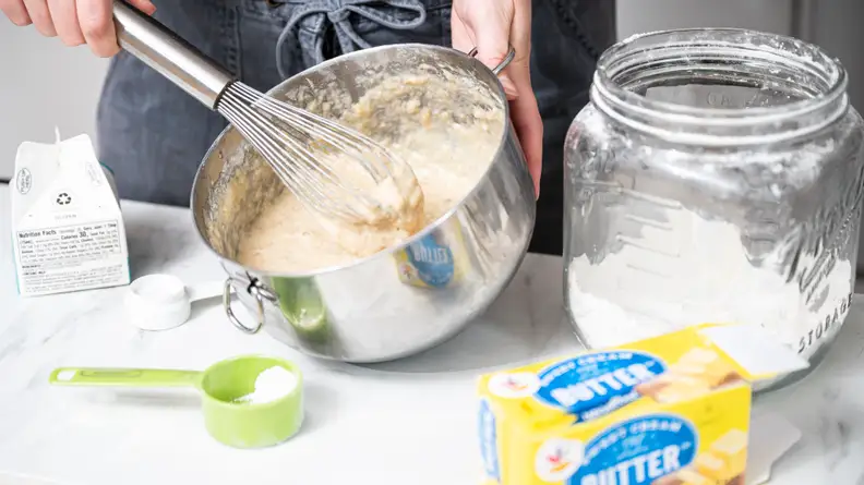 A person uses a whisk to stir cake batter in a metal mixing bowl.