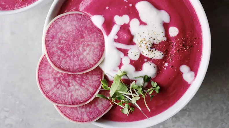 A bowl of bright pink soup garnished with radishes and white sauce